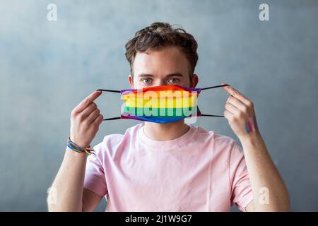 Transgender-Mann, der die Kamera anschaut, während er im hellen Raum eine Schutzmaske mit einem Regenbogen-LGBT-Symbol auf grauem Hintergrund aufsetzt Stockfoto