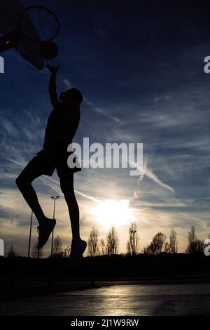 Ganzkörper-Silhouette eines gesichtslosen aktiven Sportlers in Sportbekleidung, der mit Ball springt, während er abends auf dem Spielplatz Basketball spielt Stockfoto