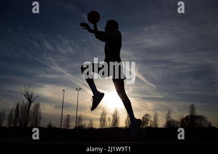 Ganzkörper-Silhouette eines gesichtslosen aktiven Sportlers in Sportbekleidung, der mit Ball springt, während er abends auf dem Spielplatz Basketball spielt Stockfoto