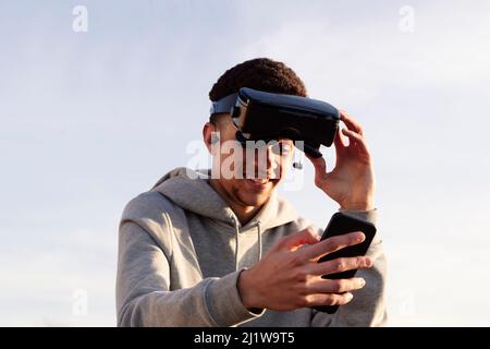 Positiver hispanischer Mann in moderner VR-Brille, der am sonnigen Tag in der Stadt die virtuelle Realität gegen den wolkenlosen Himmel erkundet Stockfoto