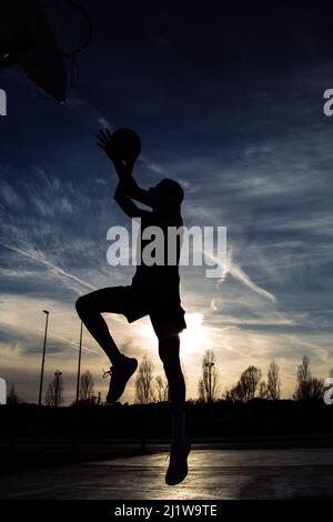 Ganzkörper-Silhouette eines gesichtslosen aktiven Sportlers in Sportbekleidung, der mit Ball springt, während er abends auf dem Spielplatz Basketball spielt Stockfoto