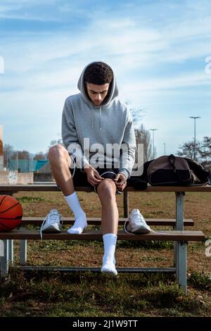 Ganzkörper des hispanischen Sportlers im aktiven Ohr, der sich auf das Basketballtraining vorbereitet, während er am Sommertag auf einer Holzbank im Park sitzt Stockfoto