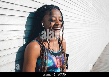 Fröhliche afroamerikanische Frau mit langen Dreadlocks, die aktive Ohrhörer für das Outdoor-Fitnesstraining und Musik in den Kopfhörern tragen Stockfoto