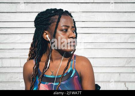 Seriöse afroamerikanische Frau mit langen Dreadlocks, die aktive Ohrstöpsel für das Fitnesstraining im Freien trägt und Musik in den Kopfhörern hört, während sie schaut Stockfoto