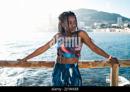 Nachdenkliche afroamerikanische Hündin mit langen Dreadlocks, die aktive Kleidung tragen und beim Fitnesstraining in der Nähe eines Holzzauns am Meer stehen und einen Blick darauf haben Stockfoto