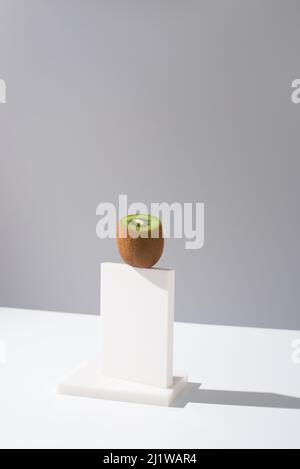 Schneiden Sie ungeschälte frische Kiwi auf weißem Ständer auf dem Tisch vor grauem Hintergrund im hellen Studio platziert Stockfoto