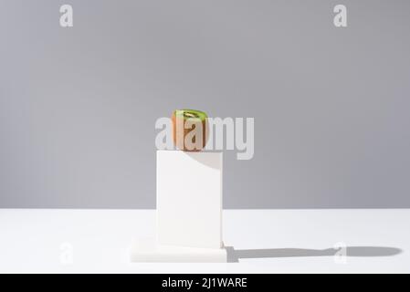 Schneiden Sie ungeschälte frische Kiwi auf weißem Ständer auf dem Tisch vor grauem Hintergrund im hellen Studio platziert Stockfoto