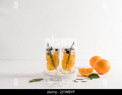 Zwei Gläser Wasser mit Orange und Blaubeeren, Rosmarinzweigen auf einem weißen Tisch gegenüber einer weißen Ziegelwand und reifen Früchten Stockfoto