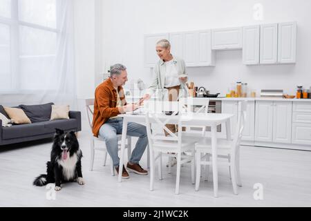 Lächelnde ältere Frau hält Teller in der Nähe des Mannes und Border Collie Hund in der Küche Stockfoto