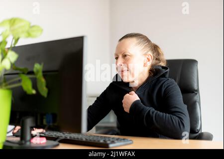 Lächelnde Frau mittleren Alters, die am Tisch sitzt und Computer in einem kleinen Heimbüro benutzt. Stockfoto
