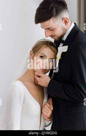 Ein stilvoller Bräutigam süß umarmt eine Braut in einem Brautkleid. Das Brautpaar umarmt sich im Zimmer und verbringt Zeit miteinander Stockfoto