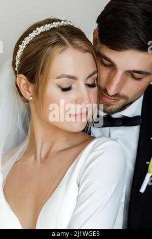 Ein stilvoller Bräutigam süß umarmt eine Braut in einem Brautkleid. Das Brautpaar umarmt sich im Zimmer und verbringt Zeit miteinander Stockfoto