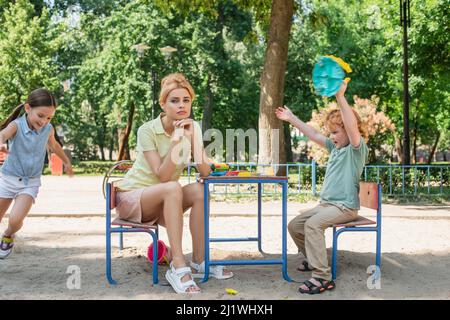 Verärgerte Frau, die neben glücklichen Kindern sitzt und Spaß auf dem Spielplatz hat Stockfoto