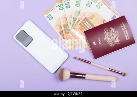 Travel Flat Lay mit weißem Smartphone, 50-Euro-Banknoten, Pass- und Make-up-Bürsten auf pastellfarbenem Hintergrund Stockfoto