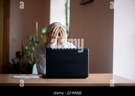 Ein müder, gebrochener, resigniert, beschäftigt älterer Mann sitzt vor einem Laptop, Computer und bedeckt sein Gesicht mit der Hand. Der traurige Senior überprüft seine Klimaanlage Stockfoto