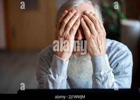 Ein müder, gebrochener, resigniert, beschäftigt älterer Mann sitzt vor einem Laptop, Computer und bedeckt sein Gesicht mit der Hand. Der traurige Senior überprüft seine Klimaanlage Stockfoto