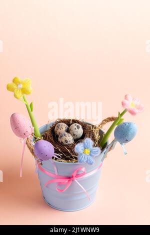 Urlaubskomposition mit kleinem Eimer mit Nest aus Wachteleiern, dekorativen Eiern, gestrickten Blumen auf pastellrosa Hintergrund. Kinder Deko-Idee für Ostern. F Stockfoto