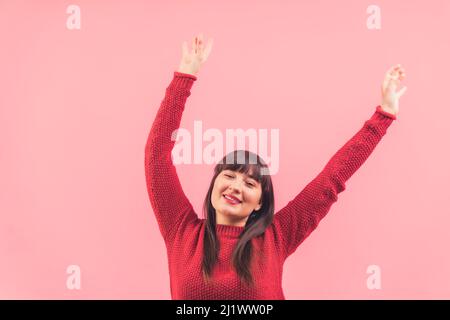 Voller Energie positive kaukasische Brünette wirft Hände in die Luft und lächelt über rosa Hintergrund. Studioaufnahme. Hochwertige Fotos Stockfoto