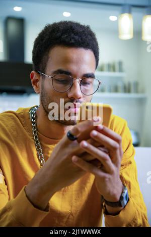 Ein junger, glücklicher Afrikaner lächelt und textete auf seinem Smartphone. Glücklicher schwarzer Mann, der zu Hause Smartphone benutzt Stockfoto