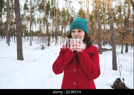 Attraktive schöne dunkelhaarige Frau in leuchtend rotem Winterwollmantel und grünem Strickhut, die Tee oder Kaffee aus einer Thermoschale trinkt und sich im Schnee ausruht Stockfoto