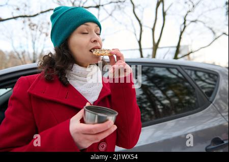 Schöne kaukasische Frau, Fahrerin Reisende Essen zum Mitnehmen, leckere gebackene Kekse, und trinken einen heißen Tee aus einem Thermobecher, stehend ne Stockfoto