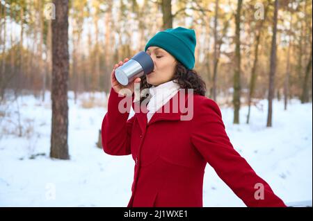 Europäische Frau in warmem roten Wollmantel und grünem Strickhut trinkt heißen Tee oder Kaffee aus einem Thermobecher aus Stahl, während sie sich in schneebedecktem Holzlant ausruht Stockfoto