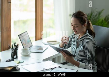 Attraktive Geschäftsfrau aus Asien, die ein digitales Tablet benutzt, während sie am Schreibtisch in einem Büro sitzt. Stockfoto