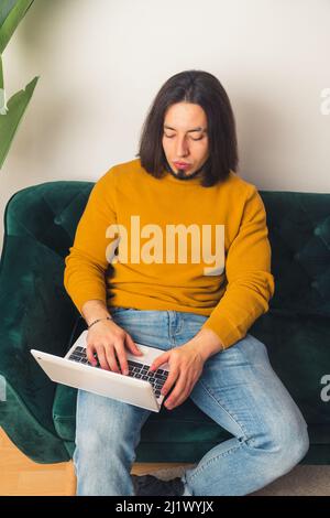 Vertikale Aufnahme eines kaukasischen bärtigen langhaarigen Mannes, der mit seinem Laptop auf einem grünen Sofa sitzt. Freizeitangebot. Hochwertige Fotos Stockfoto