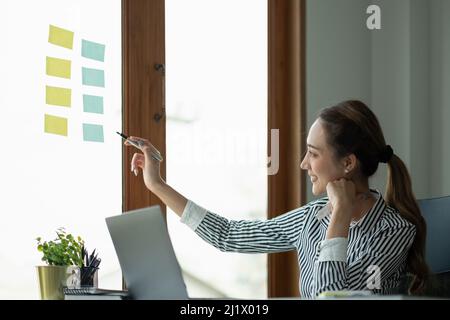 Junge kreative Frau professionelle mit Haftnotizen in Glaswand zu schreiben Strategie Business-Plan zur Entwicklung zum Erfolg wachsen. Stockfoto