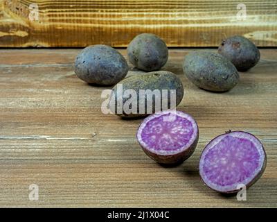 Mehrere Kartoffeln der violetten Sorte Violetta auf Holz vor einer Holzkiste, eine Kartoffel geschnitten Stockfoto