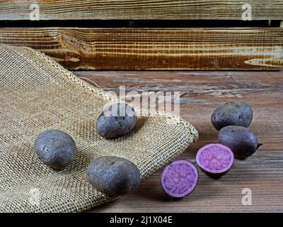 Mehrere Kartoffeln der violetten Sorte Violetta auf Holz und Sackleinen vor einer Holzkiste, eine Kartoffel geschnitten Stockfoto