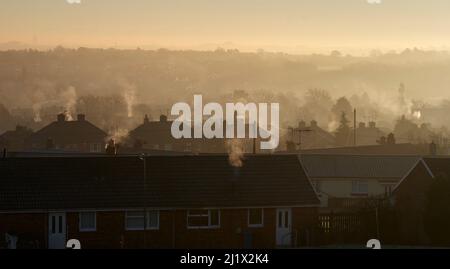 Kondensation der Abgase einer gasgefeuerten Zentralheizung, die sich im frühen Morgenlicht über den Dächern erhebt Stockfoto