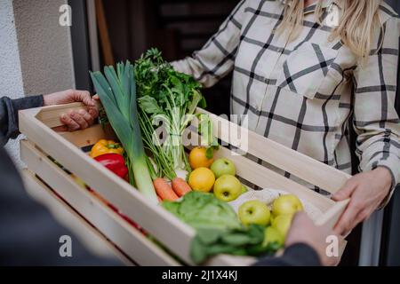 Mann, der eine Kiste mit Gemüse und Obst hält und sie der Frau, die an der Tür steht, übergibt. Stockfoto