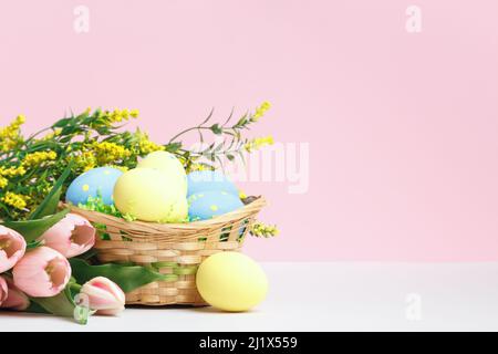 Osterkomposition. Korb mit Eiern Blumen Tulpen blühenden Ästen auf dem Tisch auf einem rosa Hintergrund. Stockfoto