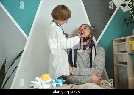 Vater und Kind spielen Klinik und Arzt, kleiner Junge Zahnarzt in medizinischen Kleid mit Stethoskop behandelt Papa Stockfoto