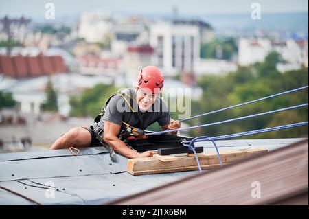 Industriebergsteiger Arbeiter in Schutzhelm mit Sicherheit Kletterseil beim Klettern Dach. Porträt eines Bergsteigers, der Sicherheitskletterausrüstung während der industriellen Hochhausarbeit verwendet. Stockfoto