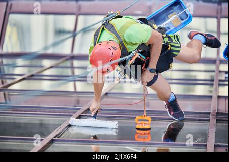 Arbeiter beim Bergsteigen waschen Glasfenster des Hochhauses, hängen an einem Kletterseil. Man Fensterputzer in Schutzhelm Reinigung Wolkenkratzer Fassade. Draufsicht. Stockfoto