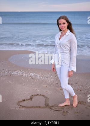 Eine junge Frau in Weiß, die am Strand steht, mit einem im Sand gezeichneten Herzen und der Kamera zuschaut. Stockfoto