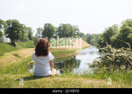 Girl Traveler sitzt auf dem grünen Gras und blickt auf den See. Rückansicht einer Frau mit lockigen Haaren, die einen schönen Blick auf das Wasser genießt. Das Konzept der Erholung in der Stille in der Natur Stockfoto