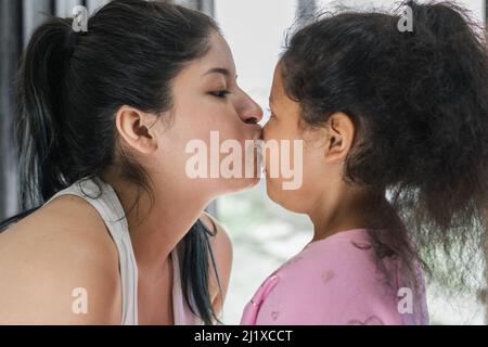 Schöne Latina Frau küsst die Nase ihrer kleinen Brünette Tochter, während sie in die Augen schaut und ihre bedingungslose Liebe zum Ausdruck bringt. Kleines Mädchen Stockfoto