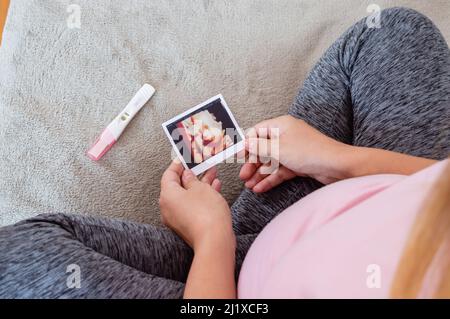 Draufsicht Schwangere, die in ihrem Zimmer sitzt und ihr Ultraschallbild ihres Babys beobachtet, mit dem positiven Schwangerschaftstest auf dem Bett Stockfoto