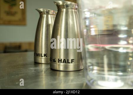 Milch und halb-halb Krüge neben einem unfokussiertem Wasserbehälter auf einer Edelstahltheke in einem Café. Stockfoto