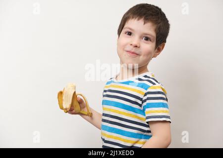 Ein 4-jähriger Vorschuljunge mit einer Banane in der Hand lächelt vor weißem Hintergrund. Stockfoto