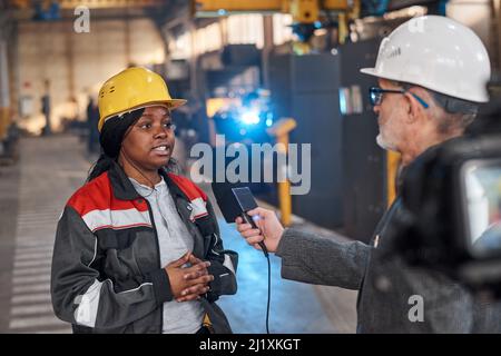 Afrikanische Arbeiterin in Uniform im Gespräch mit einer Journalistin über die letzten Nachrichten während der Reportage in der Fabrik Stockfoto