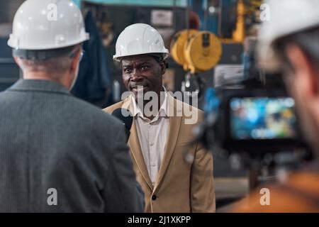 Afrikanischer selbstbewusster Ingenieur in Hardhat im Gespräch mit einem Reporter während der Online-Übertragung in der Fabrik Stockfoto