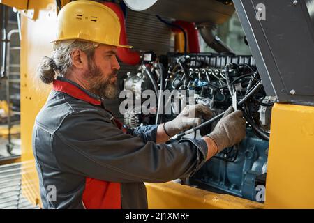 Reifer bärtiger Mechaniker in Uniform, der die Maschine untersucht und im Werk repariert Stockfoto