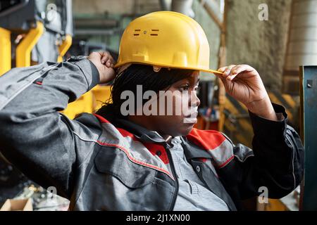 Afrikanische junge Frau in Uniform, die den Hardhut auf den Kopf stellt und ihre Arbeit in der Pflanze beginnt Stockfoto