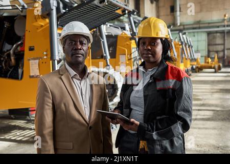 Porträt einer afrikanischen Ingenieurin mit einer Mechanikerin, die in der Fabrik steht und die Kamera anschaut, während sie online am Tablet-pc arbeitet Stockfoto