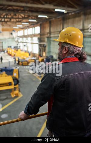 Rückansicht eines reifen Vorarbeiters im Arbeitshelm, der auf dem Balkon steht und den Produktionsprozess in der Fabrik beobachtet Stockfoto
