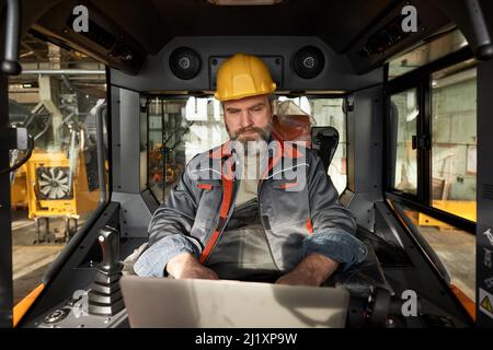 Reifer bärtiger Arbeiter in Uniform mit Laptop, während er während der Arbeiten auf der Baustelle in der Kabine der Maschine sitzt Stockfoto